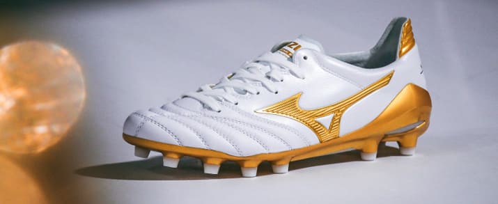 Botas de fútbol Mizuno, sobre cesped aritificial, Diseño sobrio en color blanco con pequeños detalles en dorado. Combina el estilo clásico con las necesidades del fútbol moderno.